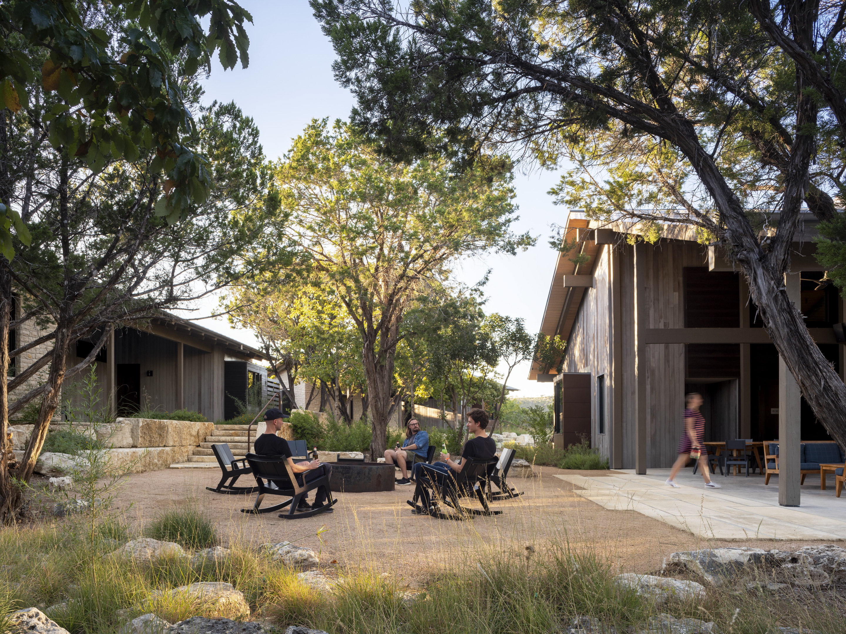 Several people sit in chairs around a fire pit in an outdoor patio area surrounded by trees and modern buildings. The serene setting's impact is enhanced as one person walks on a stone path nearby.