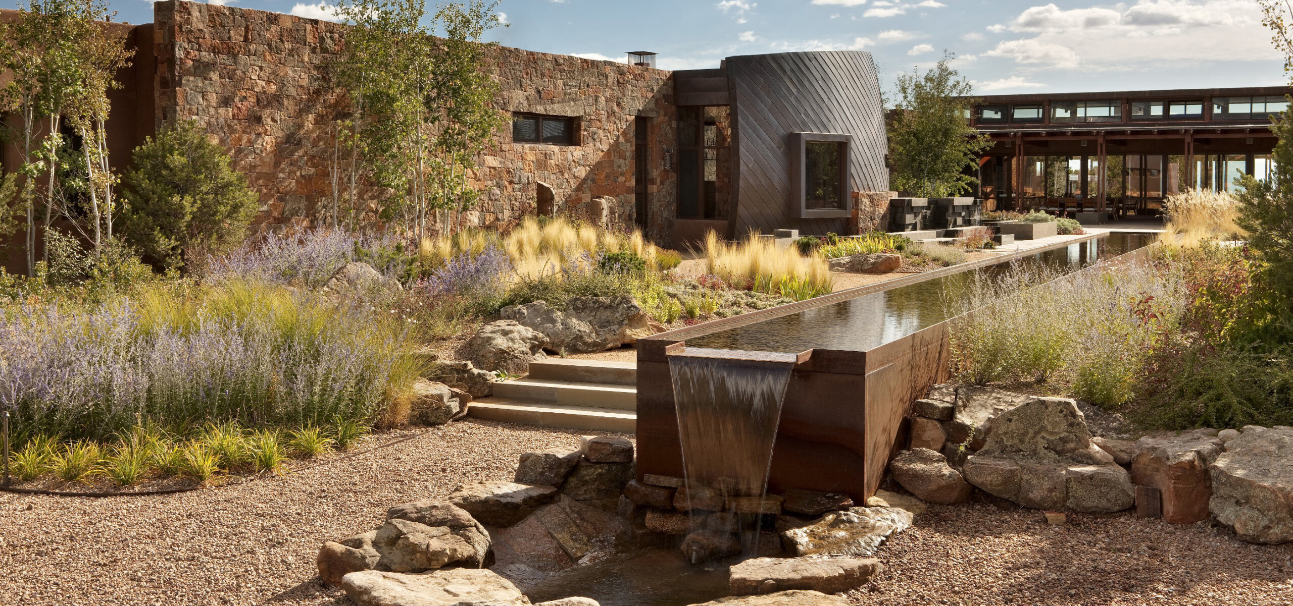 Modern architectural building with rustic stone walls, surrounded by landscaped gardens featuring rocks, grasses, and a long water feature with a cascading waterfall.