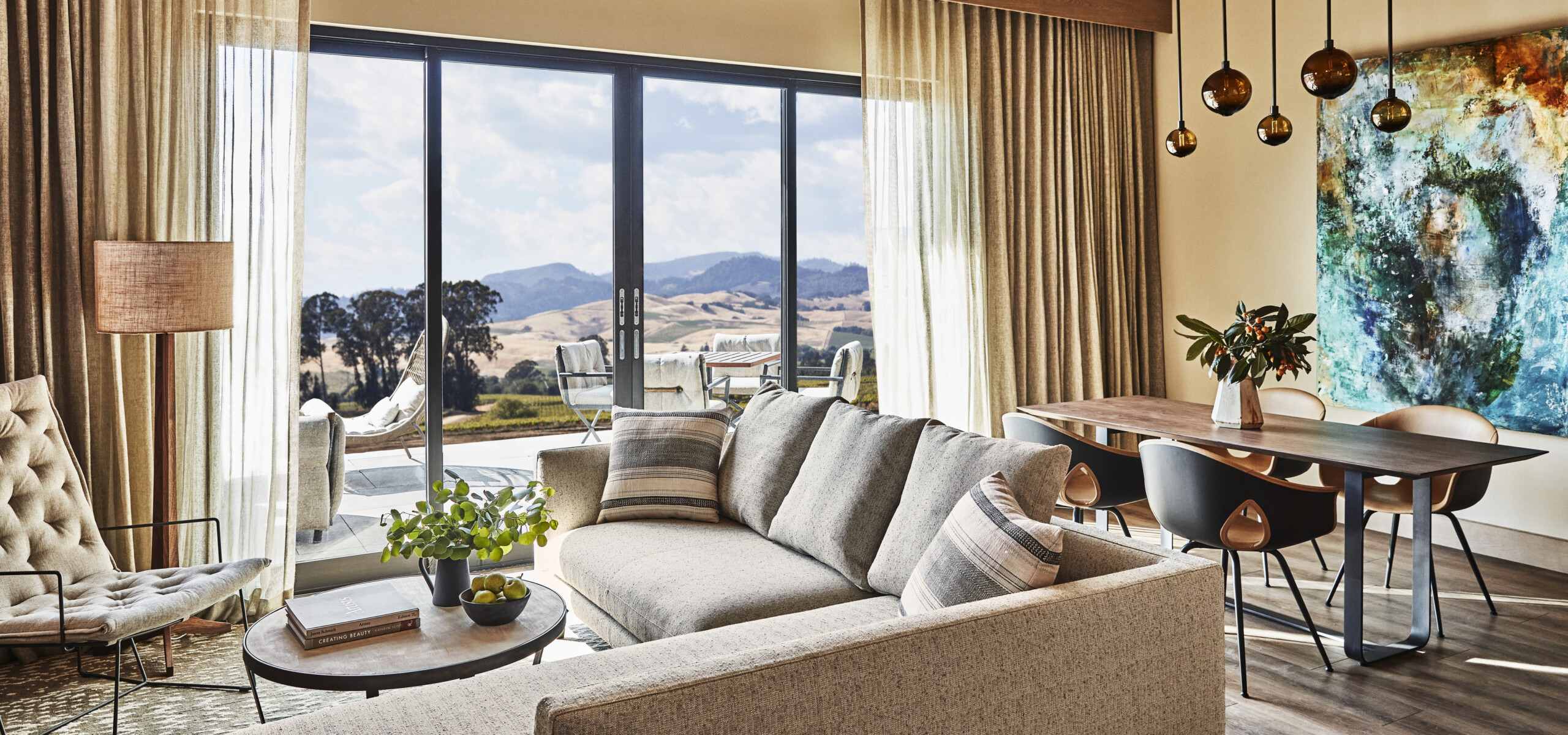 A modern living room with a large sectional sofa, coffee table, and floor lamp, featuring a dining table with black chairs, pendant lights, and a panoramic window with a countryside view.