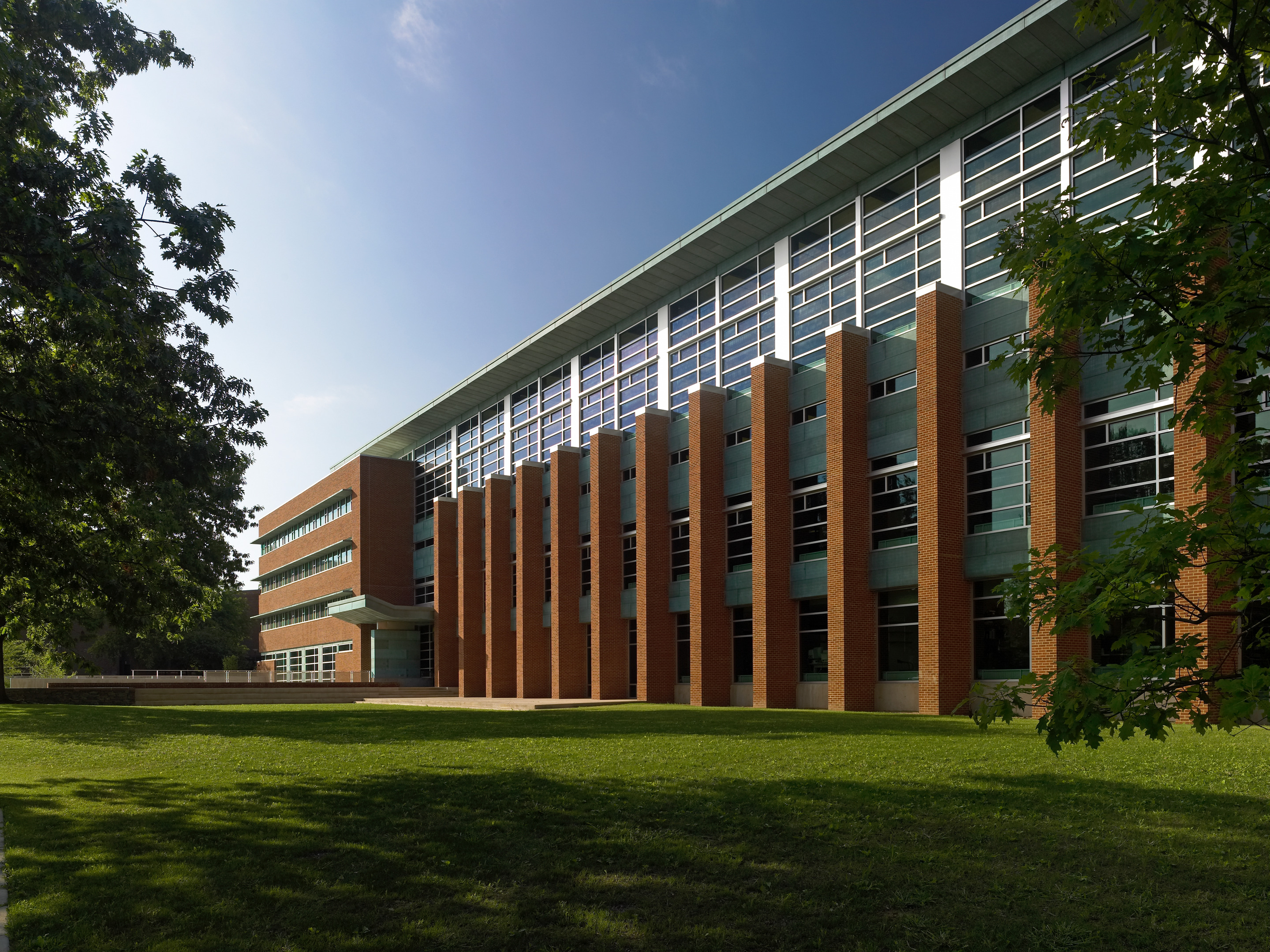 A modern, multi-story building with large windows and brick columns is surrounded by green grass and trees on a clear day, embodying the architectural excellence typical of Penn State's School of Architecture.