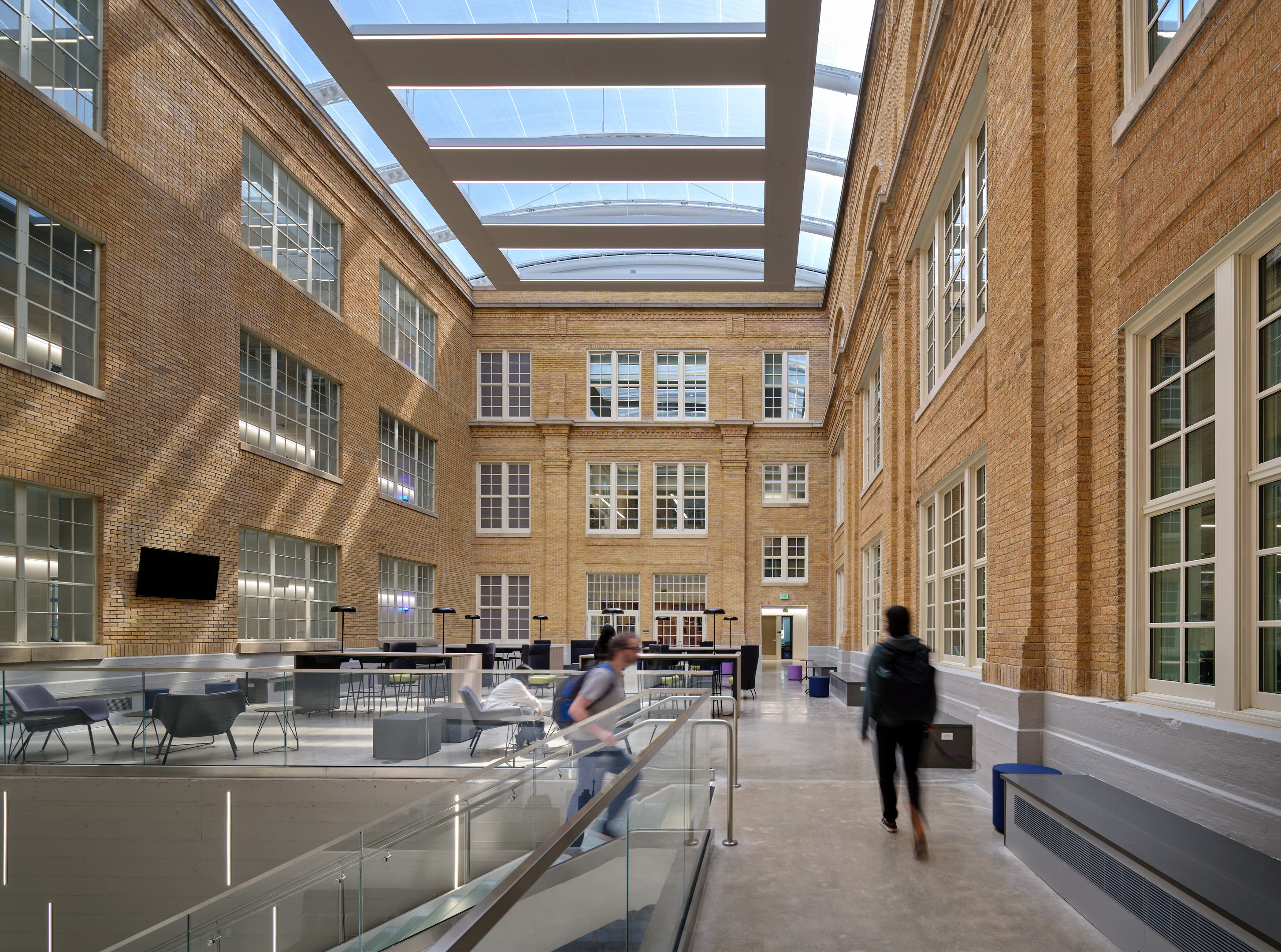 Interior of a large, modern atrium with brick walls, large windows, and a glass ceiling at Austin Community College's Rio Grande campus, featuring seating areas, a railing, and two people walking.