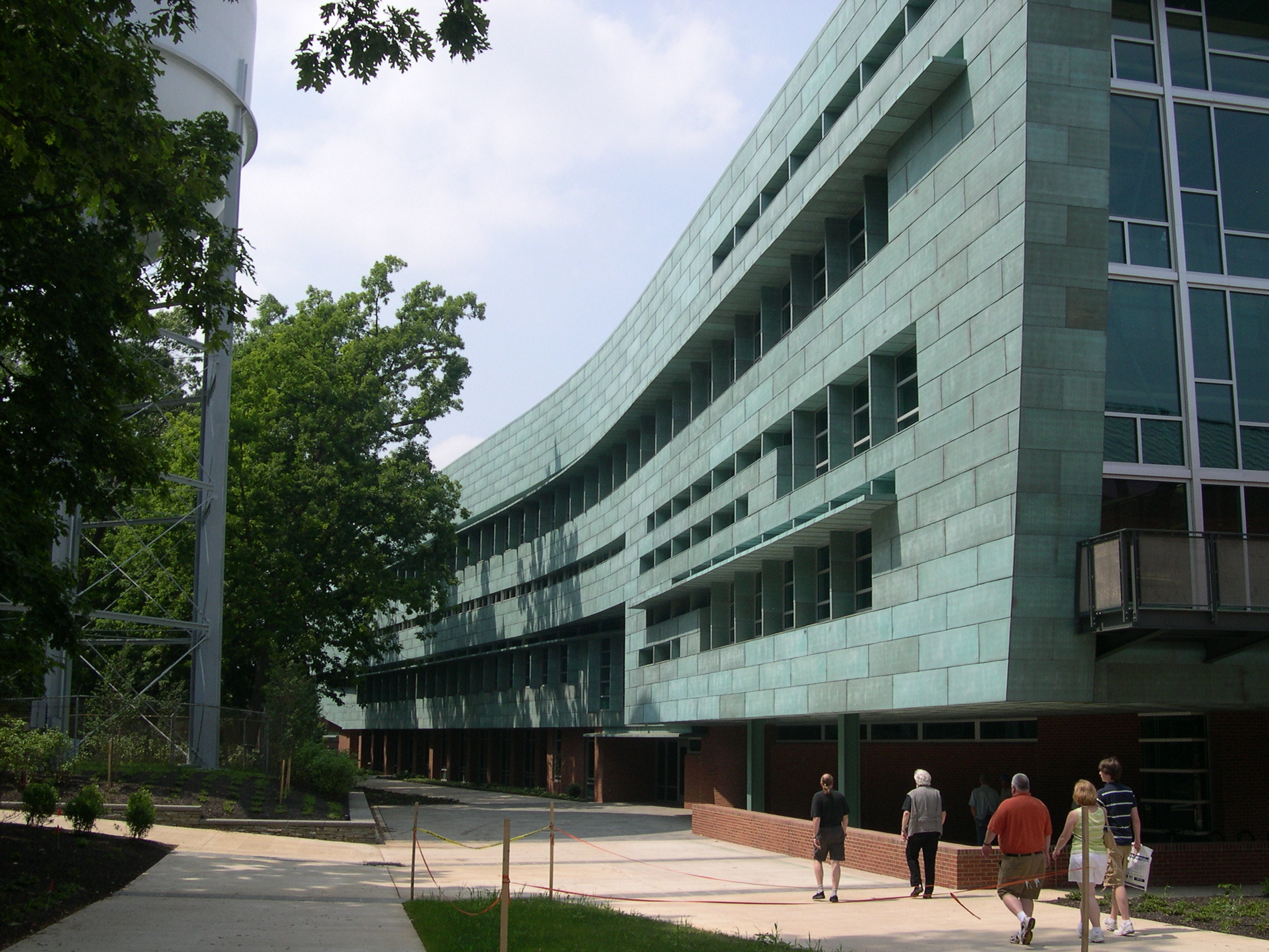 Four people walk towards a modern building with greenish exterior walls, large windows, and a wavy design, next to a white water tower and surrounded by trees. The scene exudes the innovative spirit of Penn State's School of Architecture (SOA).