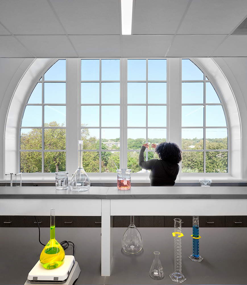 A person stands in front of large, arched windows in a laboratory at Austin Community College, holding a test tube. Various laboratory glassware and equipment are arranged on the bench. Trees and the Rio Grande landscape are visible outside.