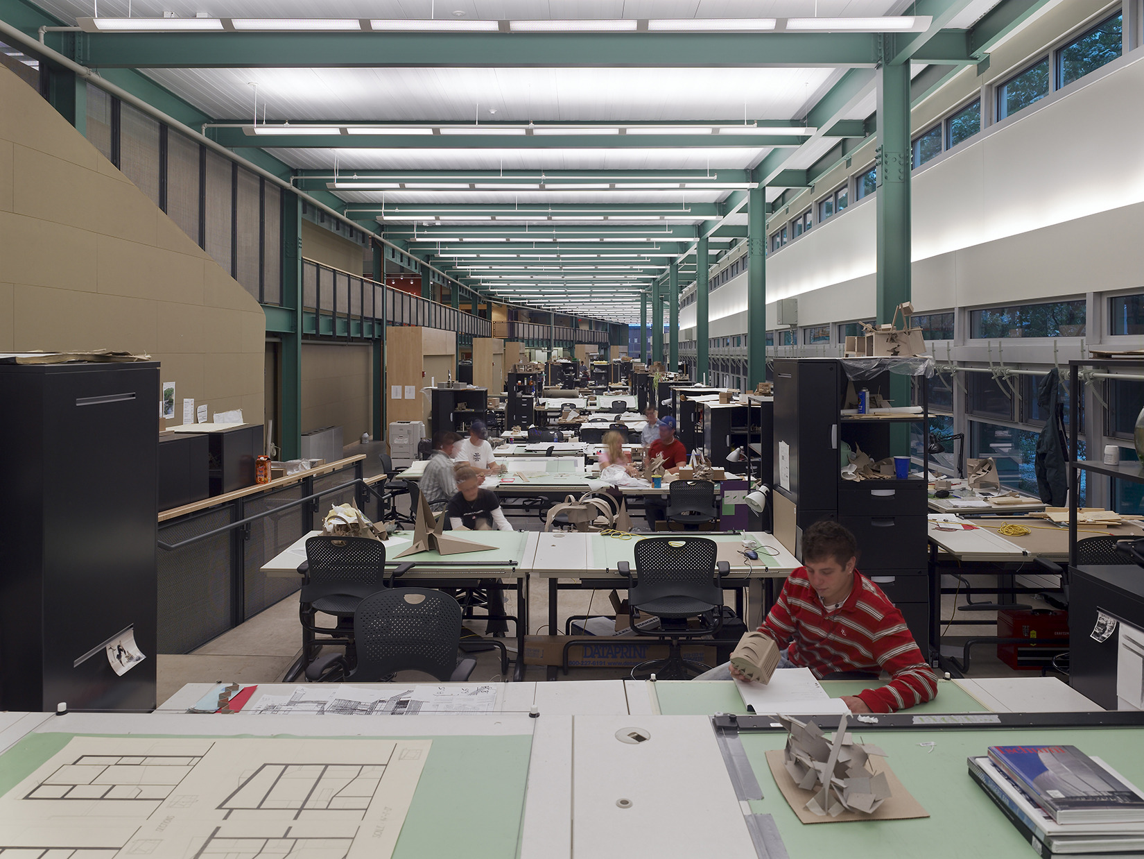 A spacious, well-lit architecture studio at Penn State, where people work at desks covered with drawings and models under a grid of ceiling lights and windows on one side.