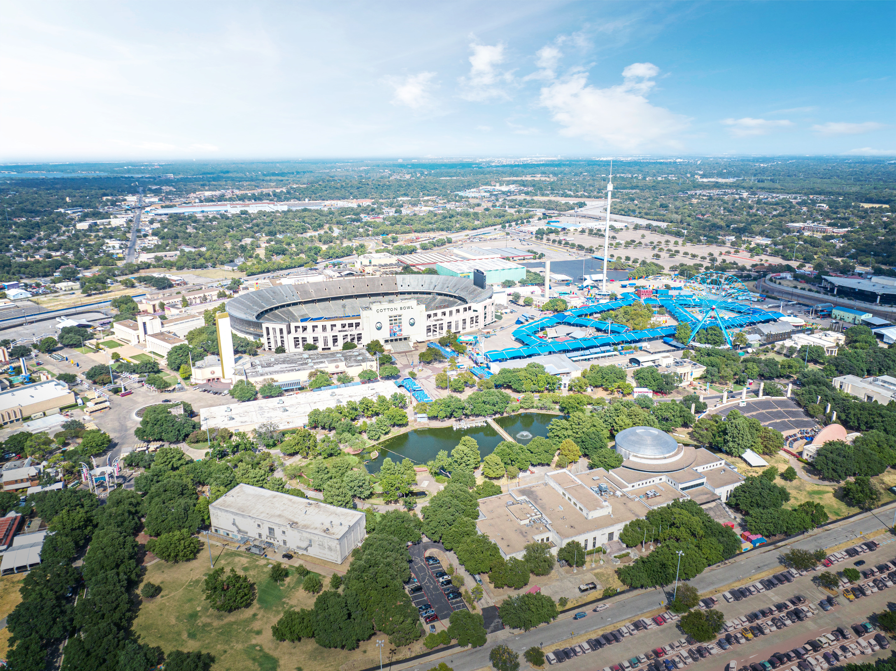 Aerial view of the Cotton Bowl stadium with adjacent fairgrounds featuring rides and booths, surrounded by green spaces and an urban landscape under a blue sky.