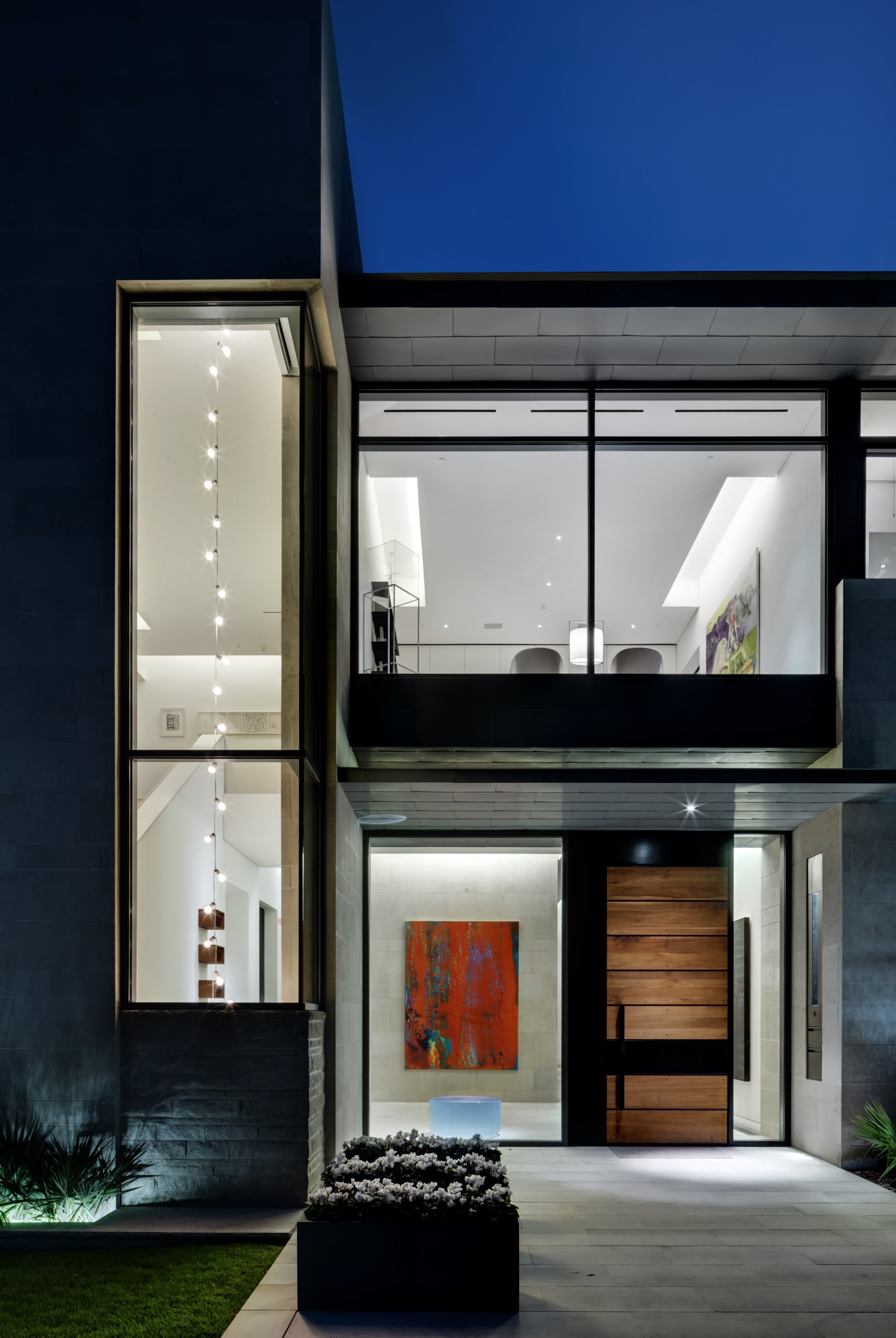 Modern residence exterior at night with large glass windows, a prominent wooden door, minimalist design, and interior artwork visible through windows.