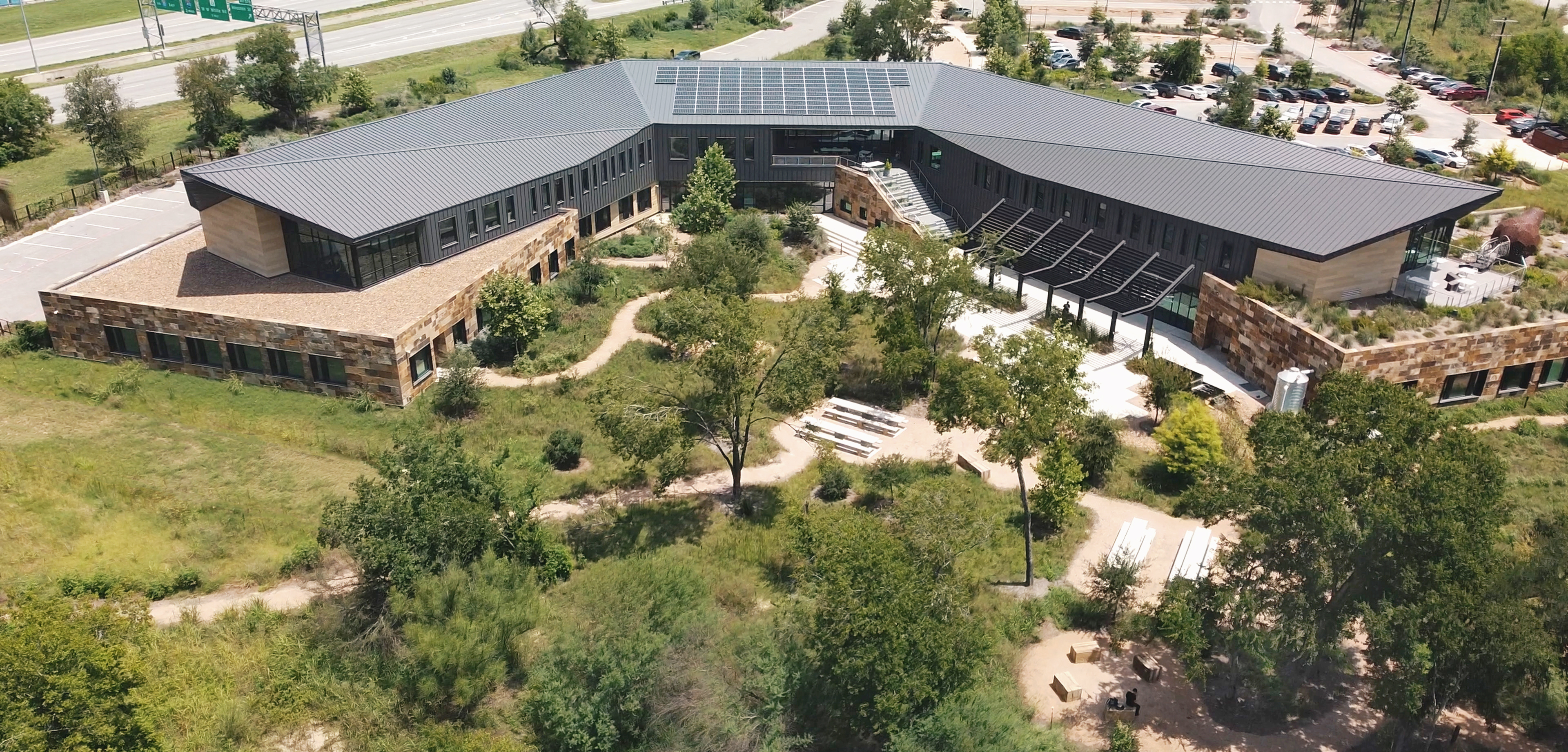 Aerial view of the Harvey E. Najim Children & Family Center surrounded by greenery, featuring solar panels on the roof and outdoor seating areas. A parking lot and a highway are visible in the background, capturing the essence of ChildSafe's commitment to sustainable family support.