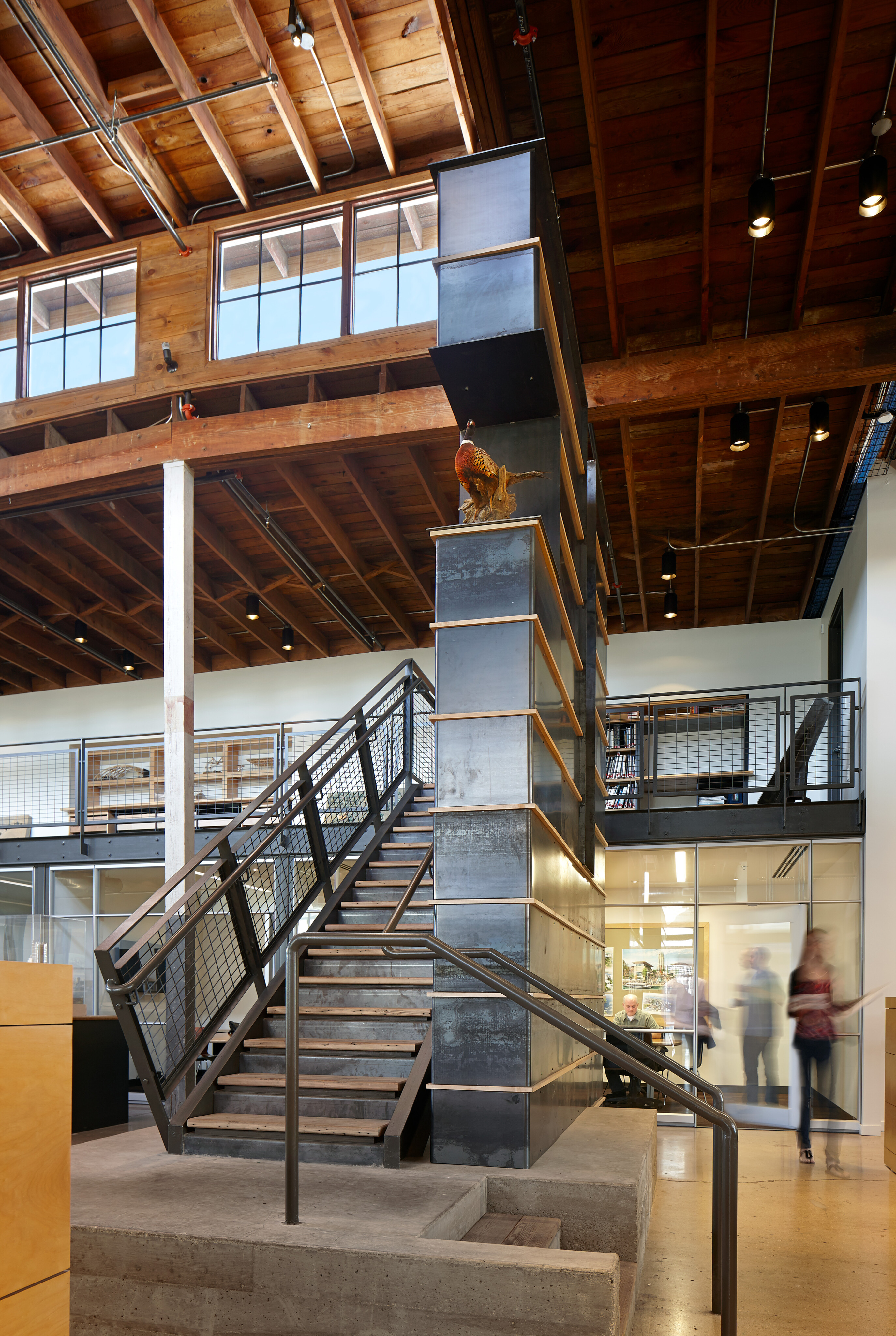 A modern office interior with a metal and wood staircase, large vertical column, glass-walled rooms, and wooden ceiling with visible beams offers a refined yet industrial vibe reminiscent of a Hughes warehouse. Two people are seen walking in the background.