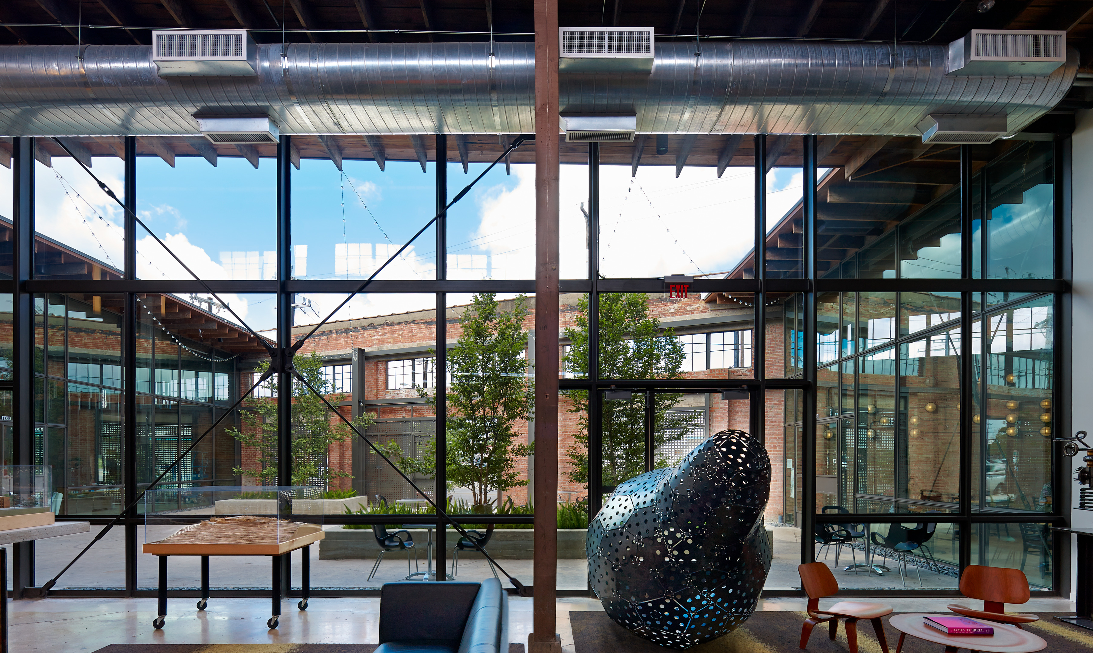 A modern, industrial-style interior with large windows revealing a courtyard. The warehouse-inspired space features ductwork, exposed beams, workstations, and a large, abstract metal sculpture.