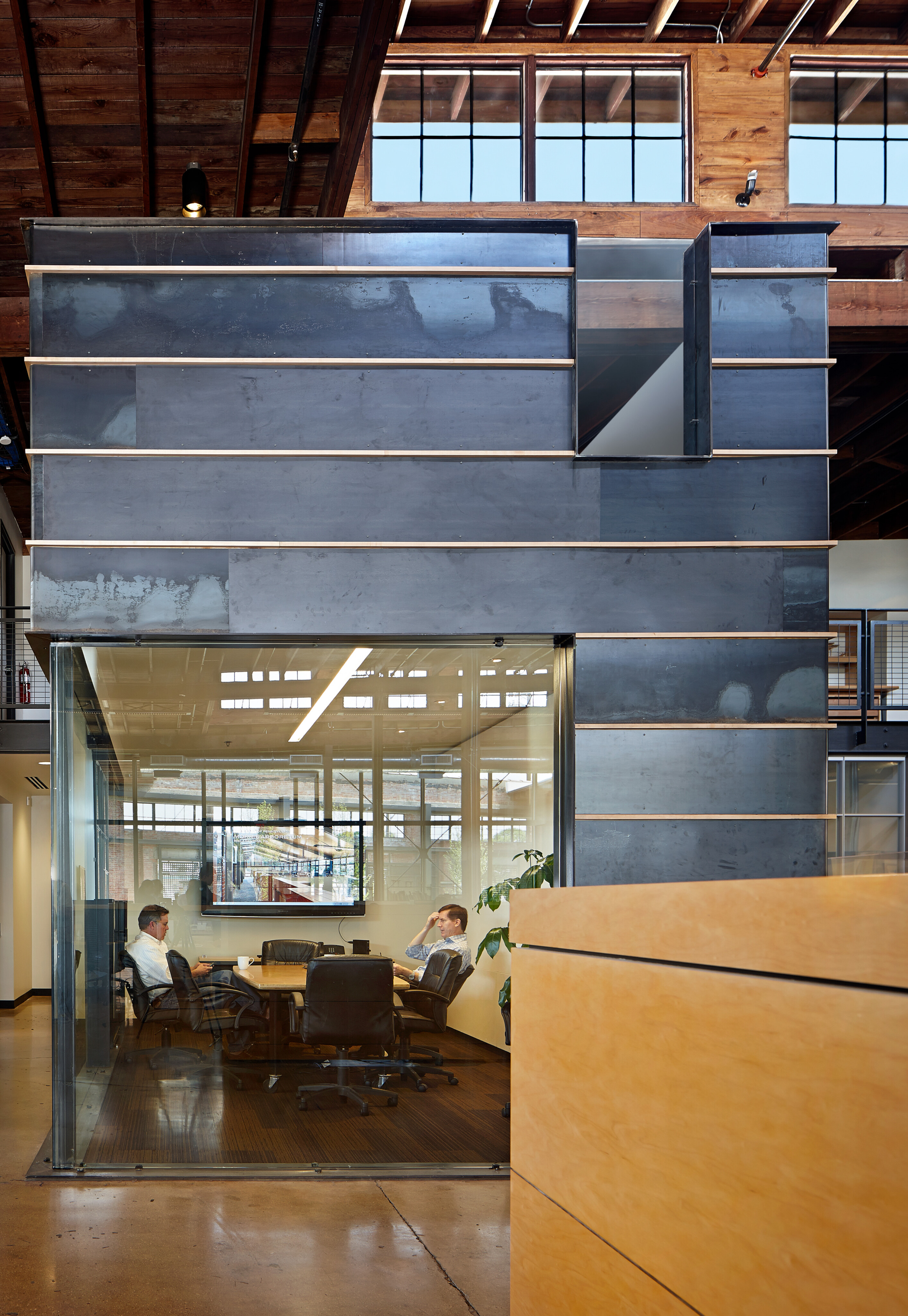 A modern office space featuring a glass-walled conference room with two people seated inside. The surrounding area, reminiscent of a sleek logistics hub, has wooden paneling, exposed ceiling beams, and a beige counter in the foreground.