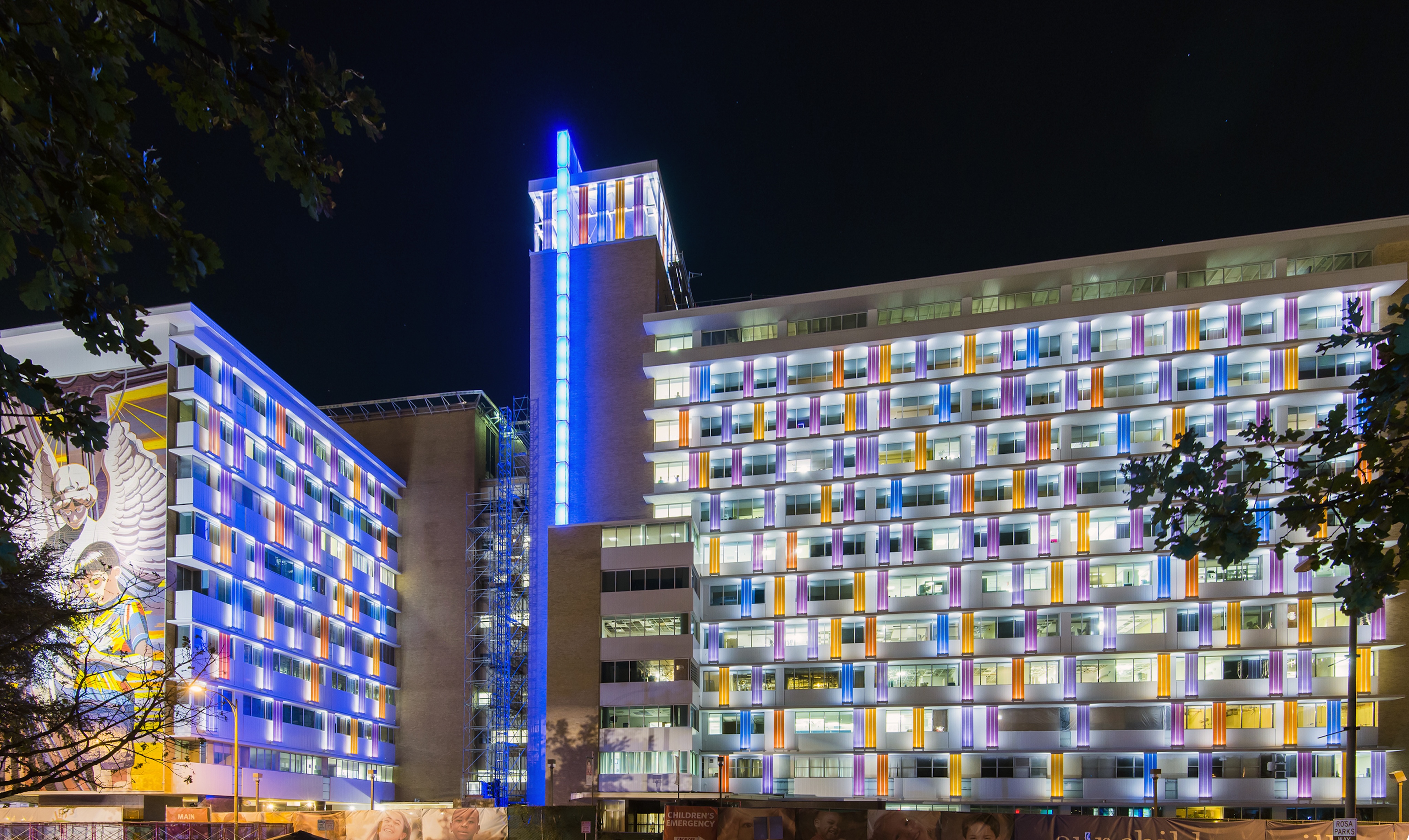 Night view of a multi-story building complex with colorful lights illuminating the windows and a central tower lit in blue. Trees partially frame the image in the foreground.