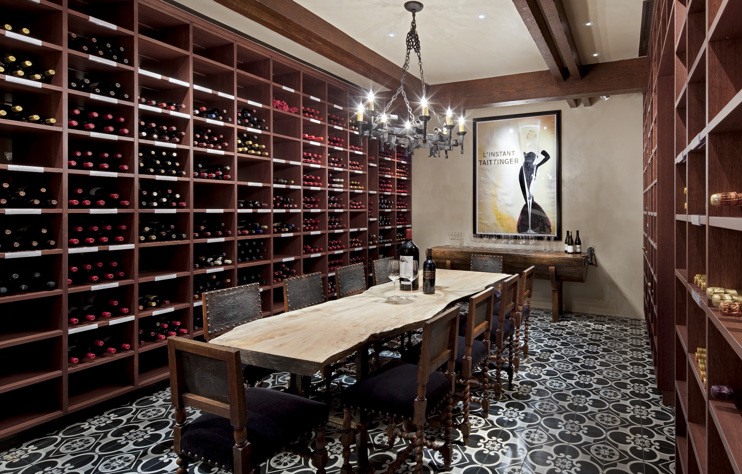 A wine cellar with a wooden table, eight chairs, and walls lined with racks of wine bottles. A chandelier hangs from the ceiling, adding an elegant touch of innovation, while a framed poster is displayed on the far wall.