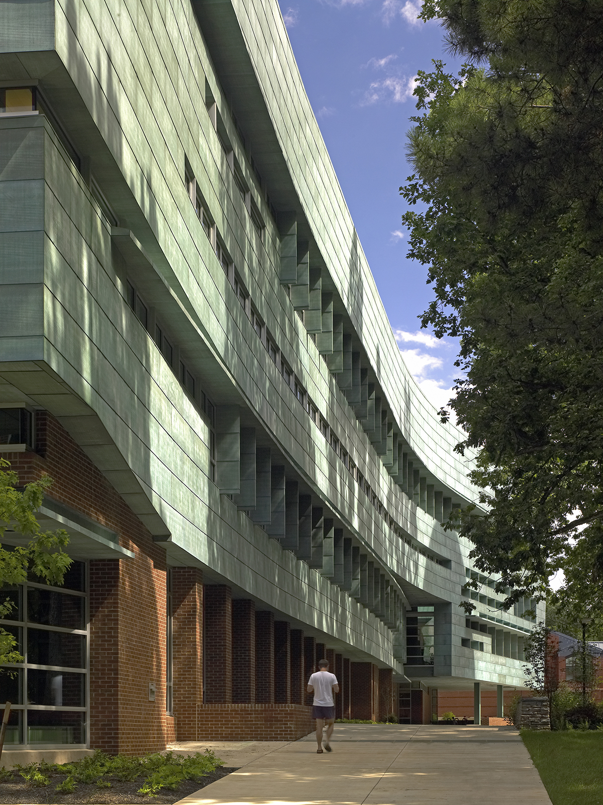 A person is walking on a path beside a large, modern building with green-tinted windows and brick accents, under a partly cloudy sky at Penn State.