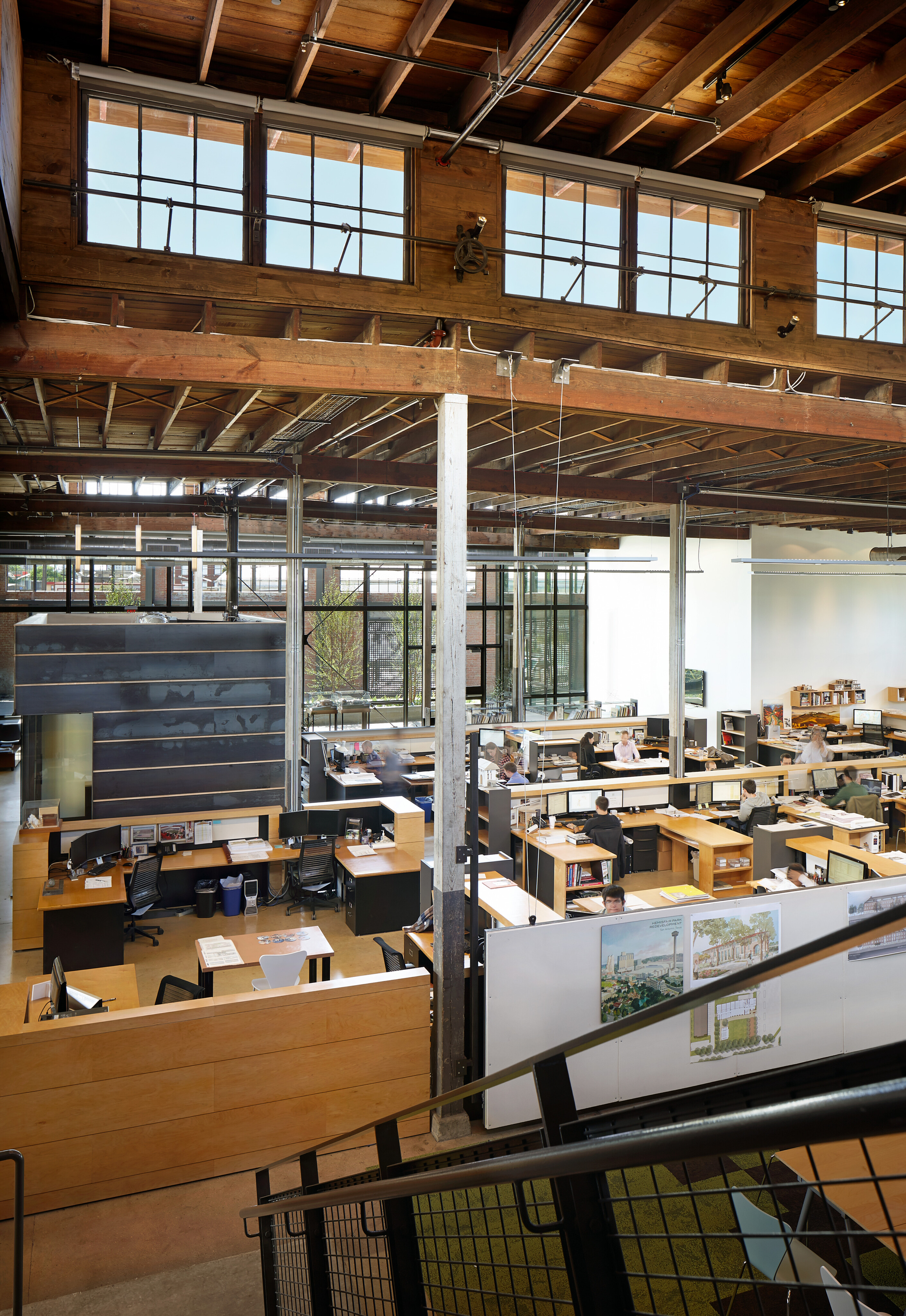 A spacious modern office with high ceilings, exposed beams, and large windows, resembling a revamped warehouse. Rows of desks and workstations are arranged in an open-plan layout, echoing the innovative spirit of Hughes.