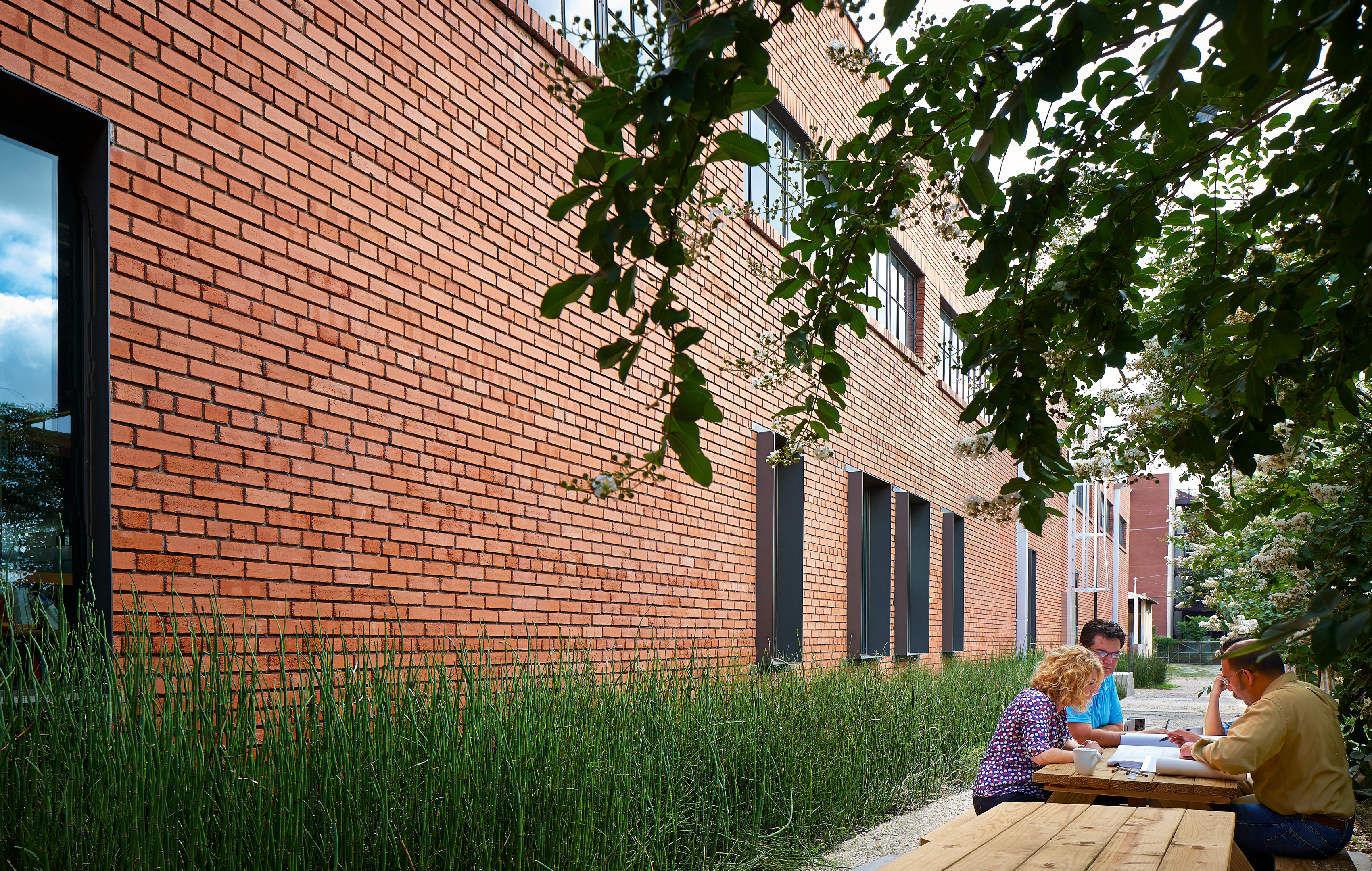 Three people sit and converse at a wooden picnic table near a large red brick building, reminiscent of an old warehouse, surrounded by greenery.