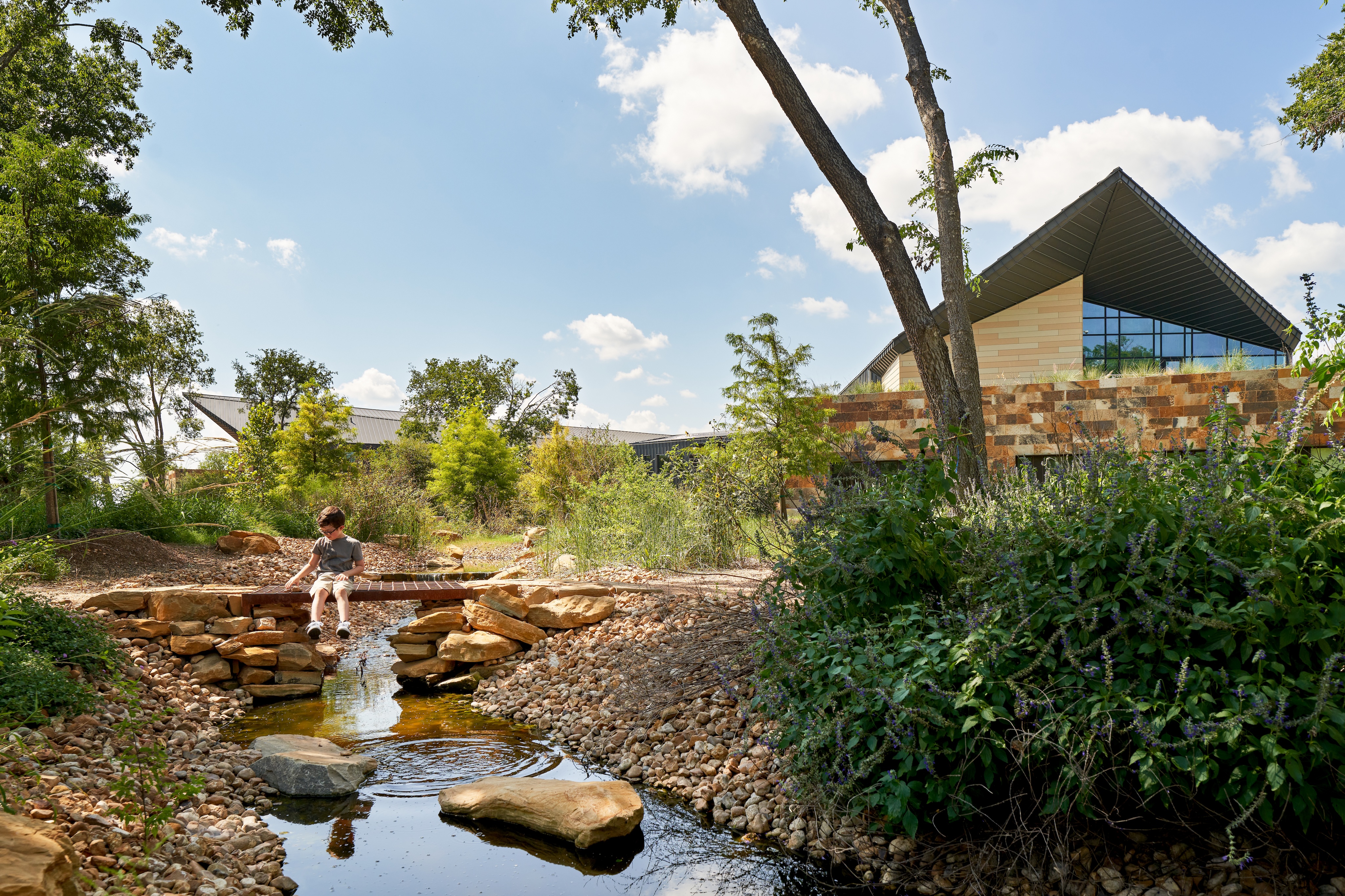 A child sits on a small bridge over a rocky stream in a park-like setting with trees and the modern, angular Harvey E. Najim Children & Family Center in the background.