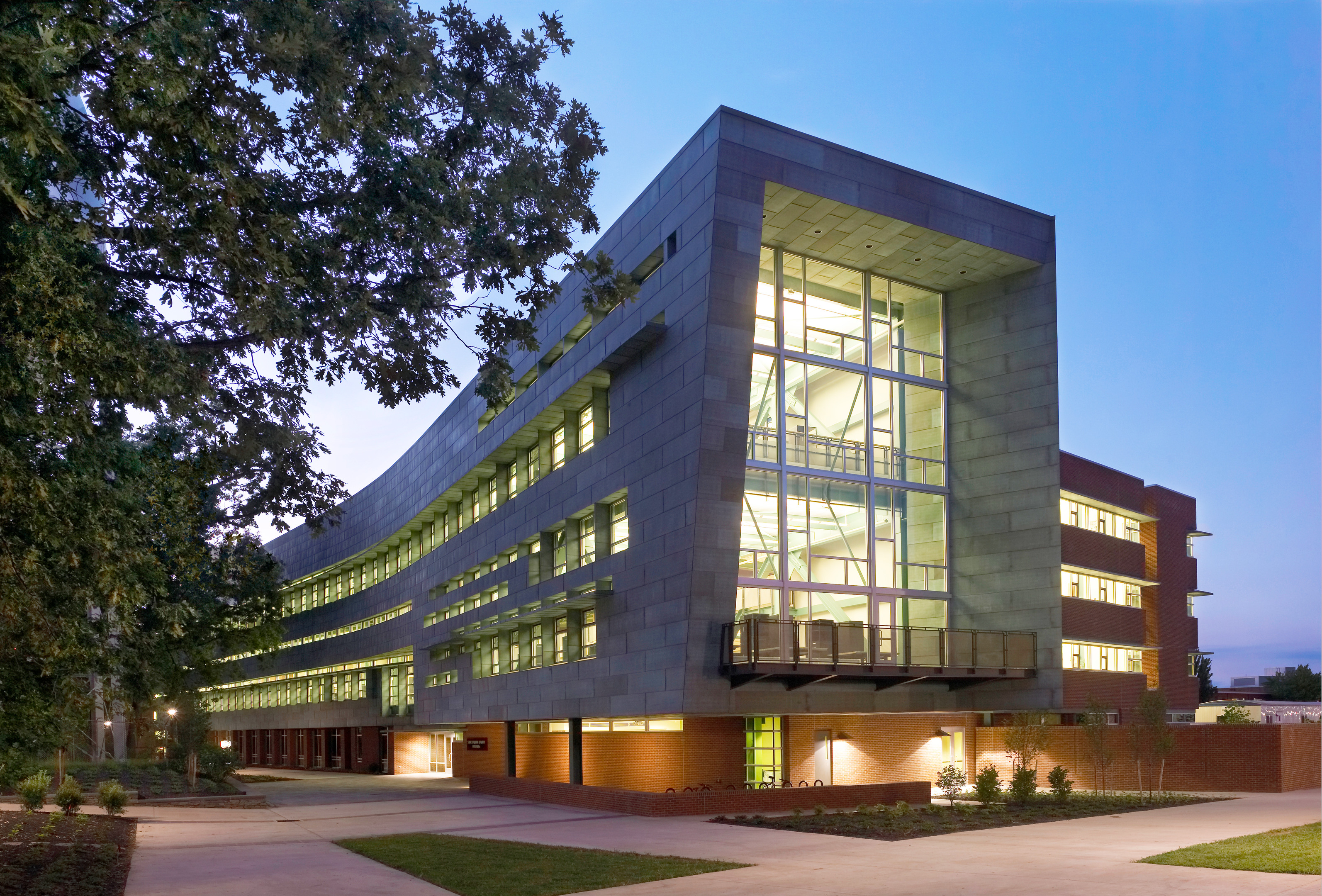 A modern, multi-story building with large windows and a unique curved design, the SOA at Penn State is surrounded by greenery and beautifully illuminated in the evening.