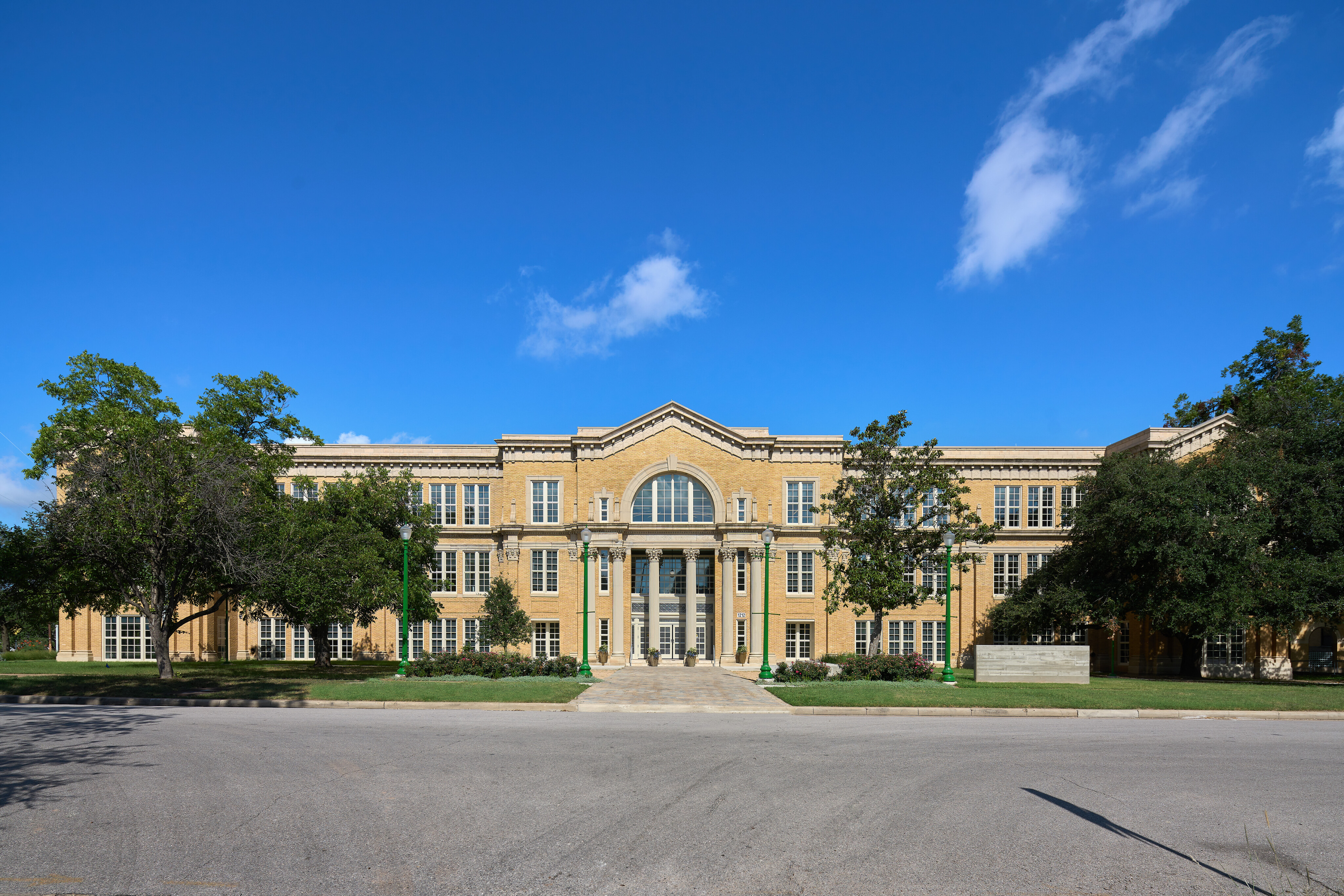 A large, historic yellow brick building with tall windows and a central arched entryway, flanked by trees, under a clear blue sky, proudly serves as the Rio Grande Campus of Austin Community College after its recent renovation.