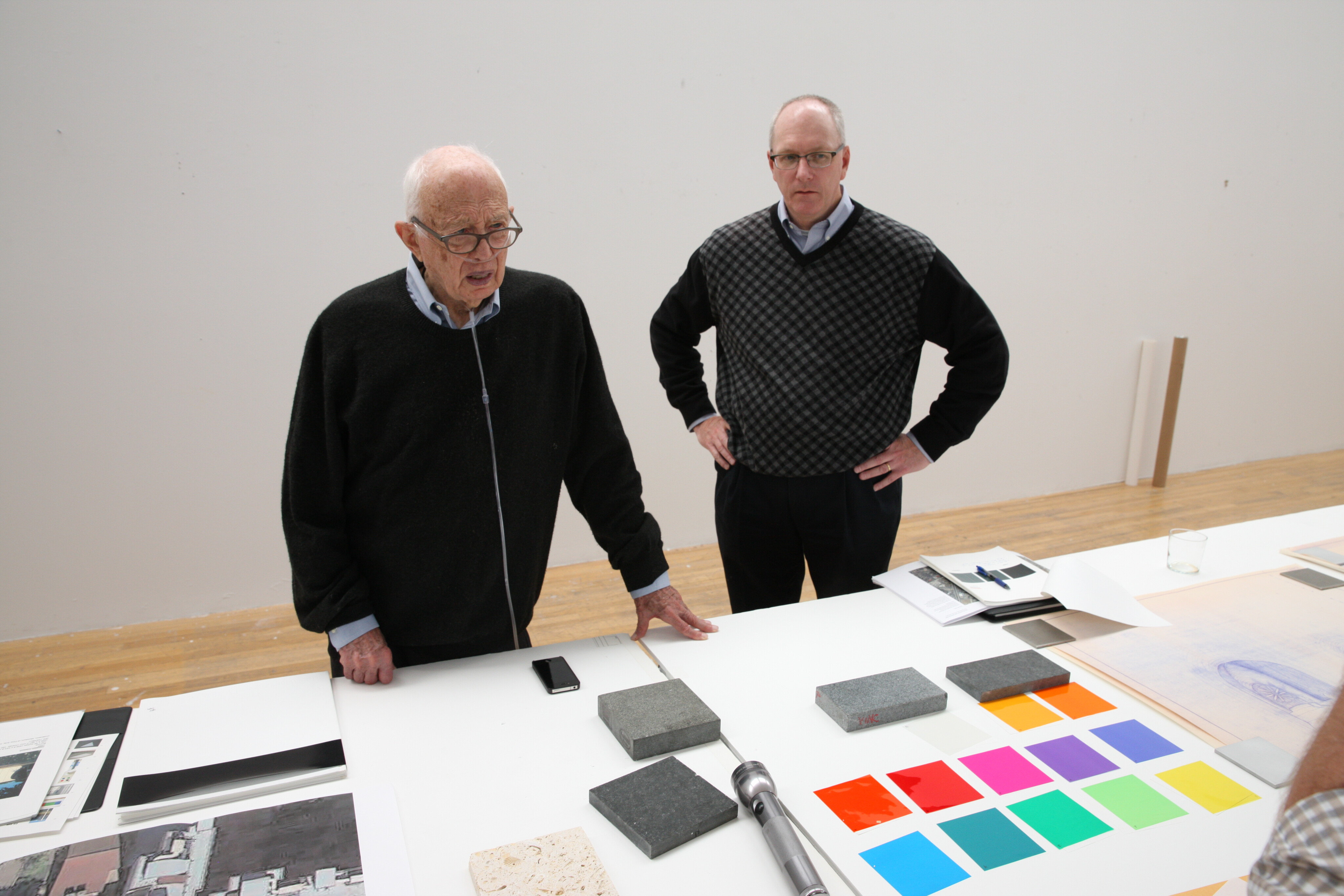 Two men stand at a table covered with various materials and color samples in a brightly lit room, reminiscent of an Ellsworth Kelly studio. Both men wear glasses and sweaters. One man speaks while the other looks towards the array of modern art supplies on the table.