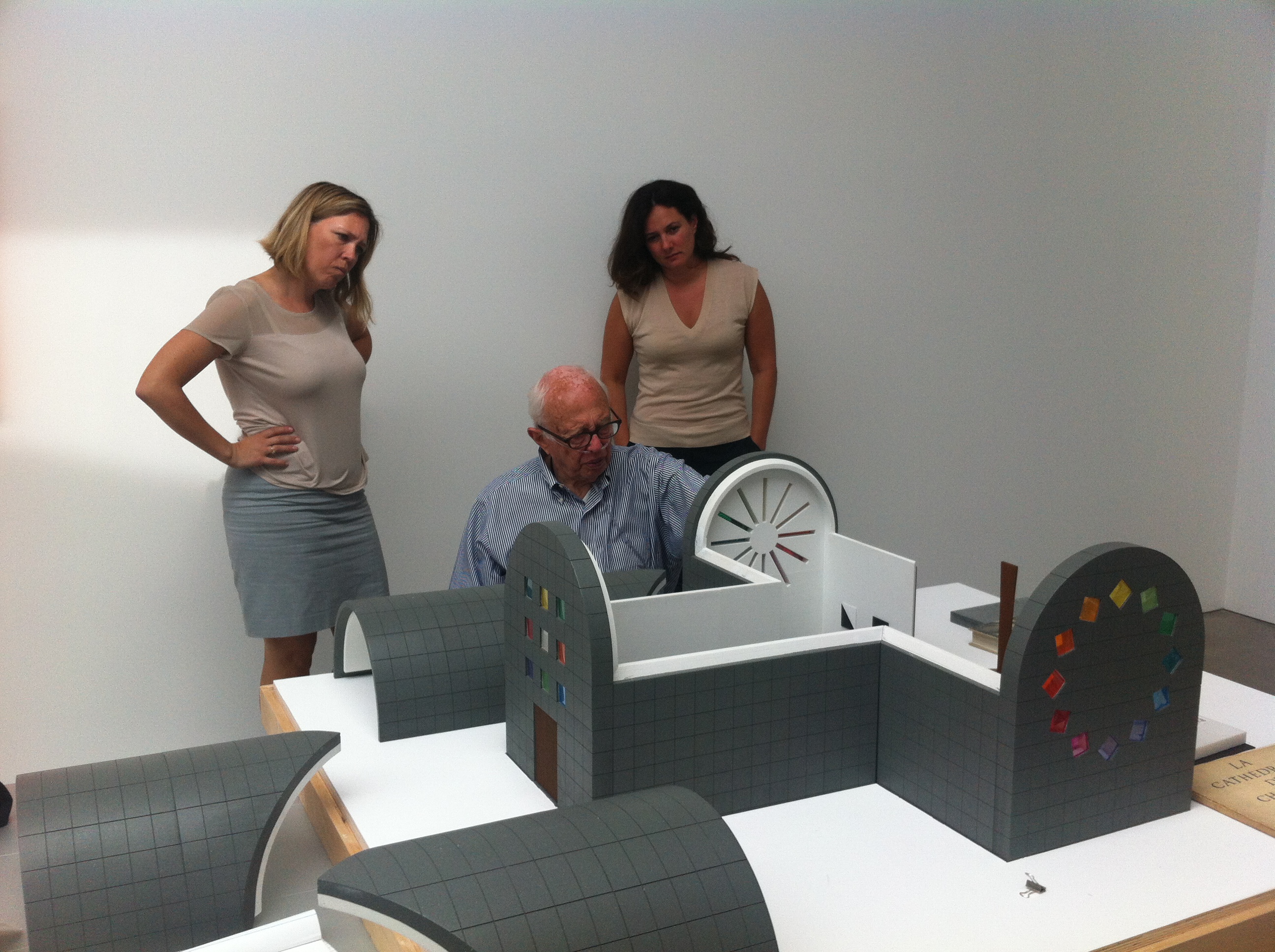 Three people are observing an artist seated at a table, working on a scale model of a building with a curved, modern design, much like the fluid forms in an Ellsworth Kelly abstract painting.