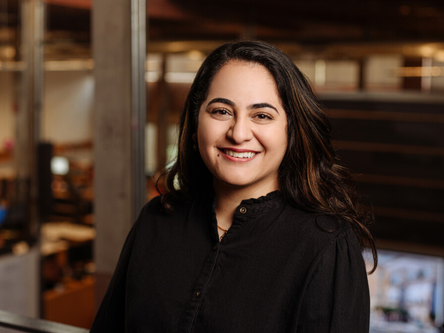 Aarti Khatter, with long dark hair, is wearing a black shirt and smiling in an indoor setting with a blurred background.