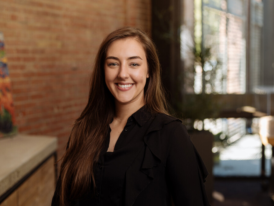 Alisha Burkman, a woman with long brown hair, is wearing a black shirt and smiling while standing indoors with brick walls and large windows in the background.