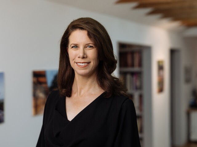 Catherine Craig, with her long dark hair, smiles while standing in a hallway with blurred shelves and pictures in the background.