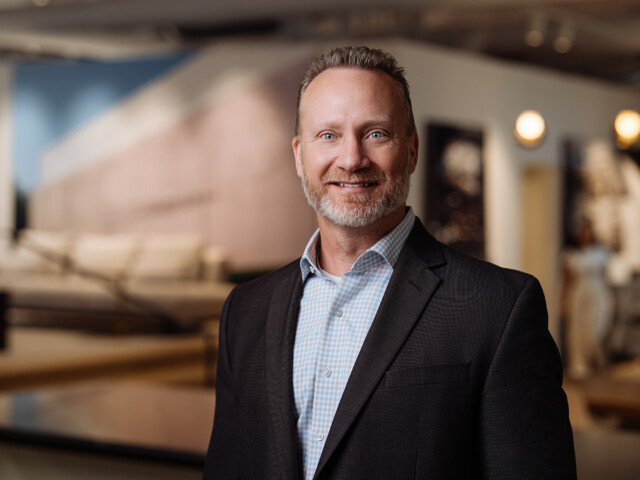 A man with a gray beard and short hair, dressed in a dark suit and light checkered shirt, stands indoors with a blurred background featuring lights and furniture. The distinguished gentleman could easily be mistaken for Chad Scheckel.