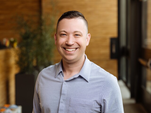 A man with short dark hair, wearing a light blue button-up shirt, smiles at the camera in an indoor setting with a blurred background; Christian Korta radiates warmth and approachability.