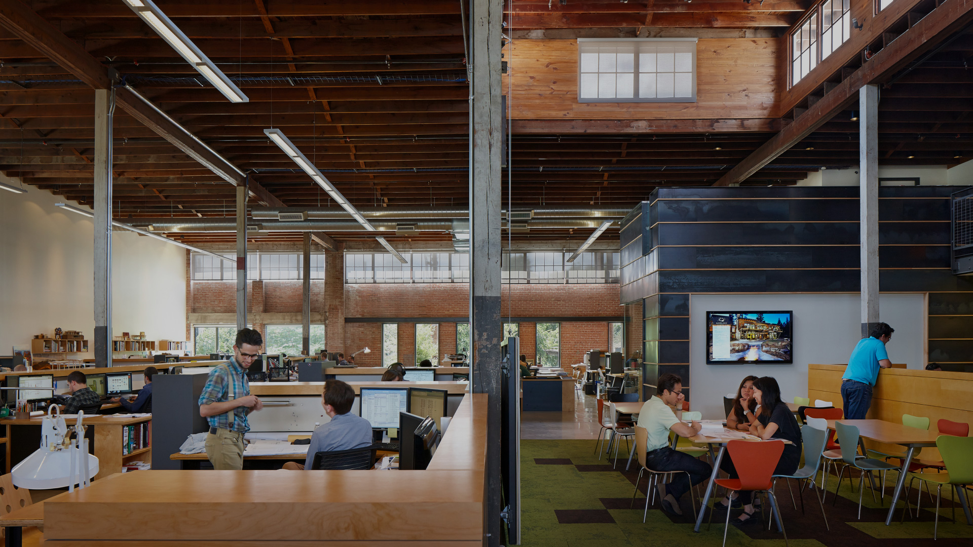 Open office space with people working at desks, a meeting happening at a table, and another group engaged in a discussion. The room features high wooden ceilings and large windows allowing natural light.