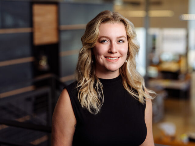 A person with long blonde hair, wearing a sleeveless black top, smiles at the camera in a modern office setting. Jenna Acord radiates confidence and warmth in this professional environment.