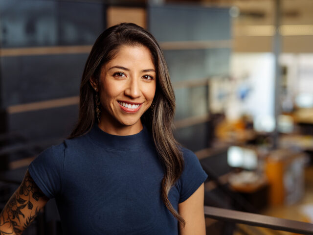 Sabrina Ortiz Luna, a woman with long hair wearing a blue top, smiles warmly in an office setting. The background shows blurred office equipment and workstations.