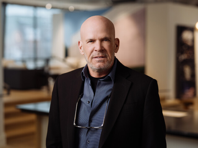 Glenn Clarke, a bald man with a neatly trimmed beard, wearing glasses on a chain, a dark blazer, and a blue shirt, stands indoors with a soft-focus background.