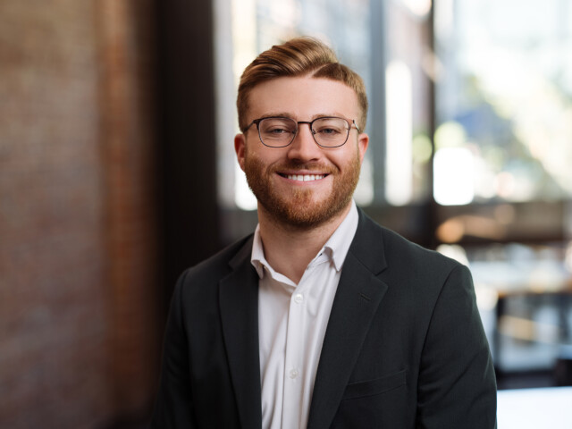 Braden King, a man with glasses and a beard, smiles while wearing a dark jacket and a white shirt. He is standing indoors, with blurred background details of bright windows and a brick wall.