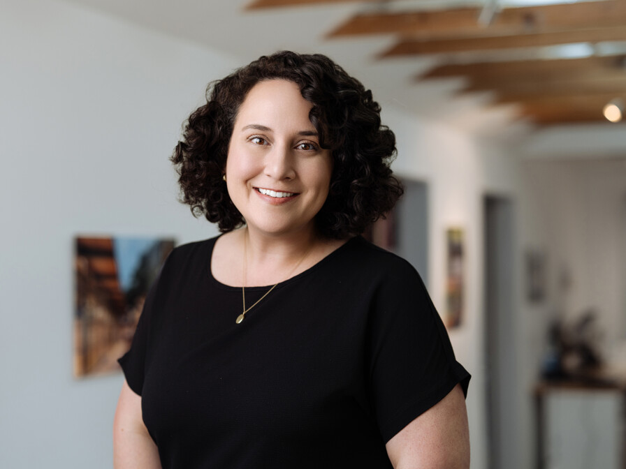 A person with curly hair, wearing a black shirt and a necklace, smiles at the camera in a well-lit room with white walls and wooden ceiling beams. Jena Hammond Nichols perfectly captures the essence of casual elegance in this candid moment.