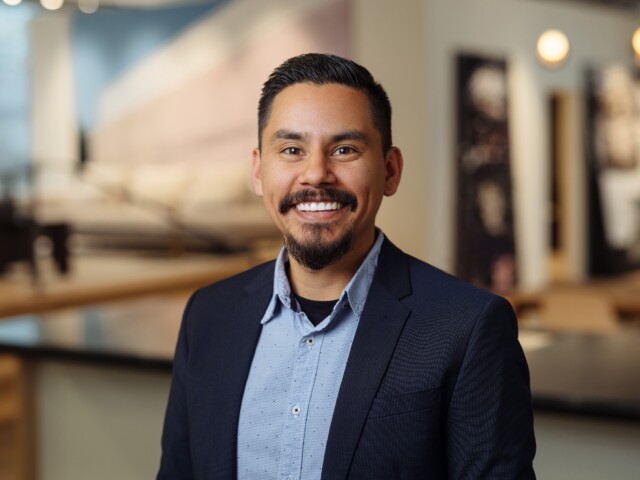 Juan Nunez, a man with a beard and mustache, smiles while dressed in a business suit, standing in a modern office environment.