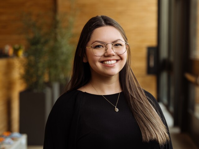 A woman with long brown hair and glasses smiles warmly. Wearing a black top, Kali Duckworth stands indoors amidst a blurred background of plants and wooden walls.