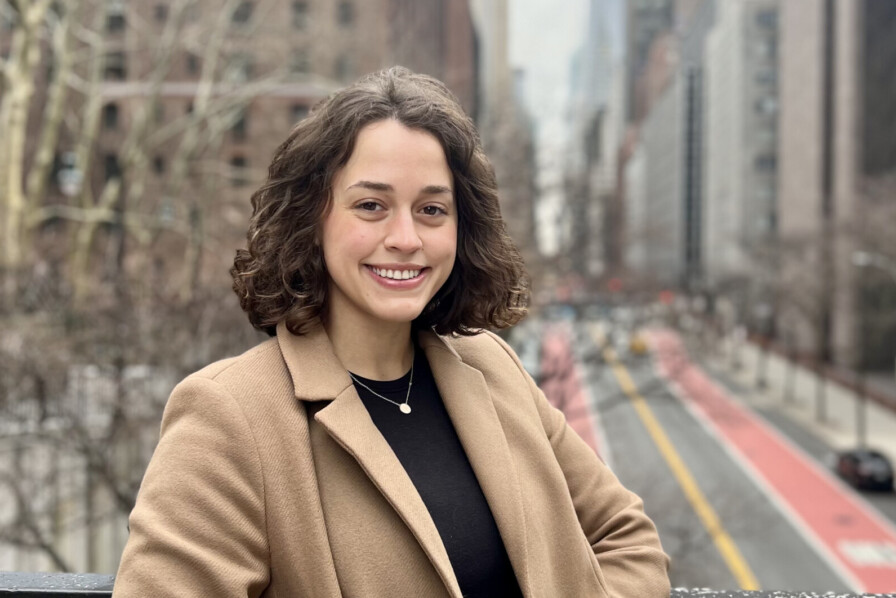 A person with curly hair and a brown coat, possibly Karina Corcuera, stands on a bridge overlooking a city street lined with tall buildings. The sky is overcast.