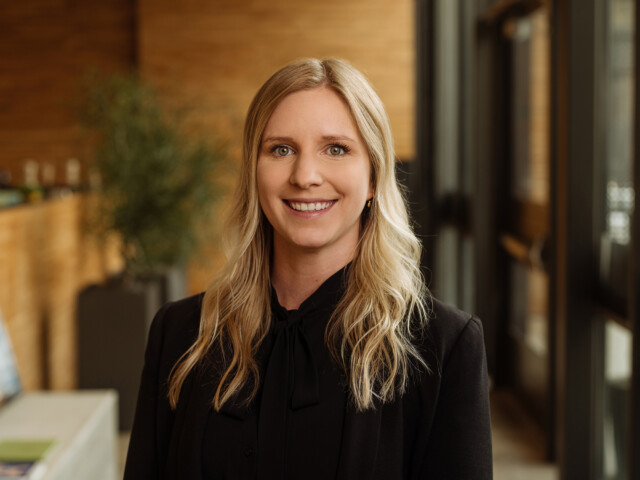 Kelsey Sletten, a woman with long blonde hair, smiles while standing in a well-lit indoor setting with wood-panelled walls, wearing a black blazer.