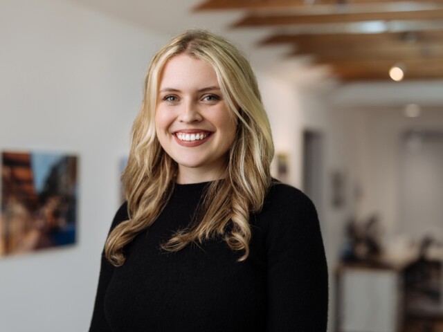 Smiling Madison Canelis with long blond hair wearing a black sweater, standing in a room with blurred background including a desk, chair, and artwork on the walls.