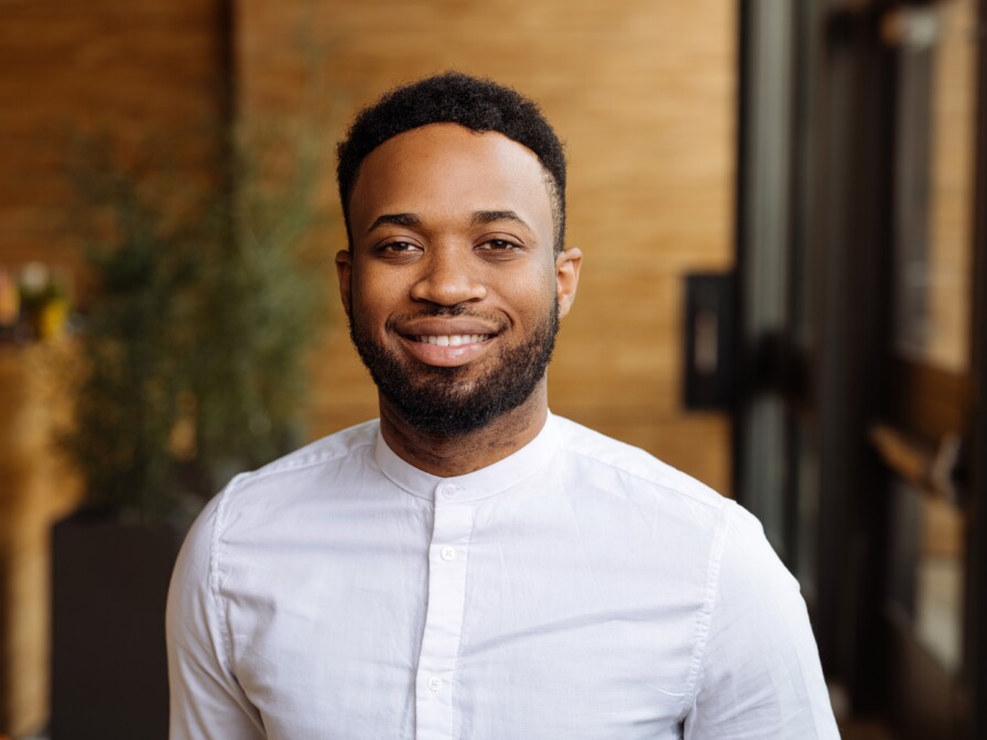 Mikah Johnson, a man with a beard and short curly hair, smiles at the camera in a warmly lit indoor setting. Dressed in a white button-up collarless shirt, this professional headshot captures his approachable demeanor perfectly.