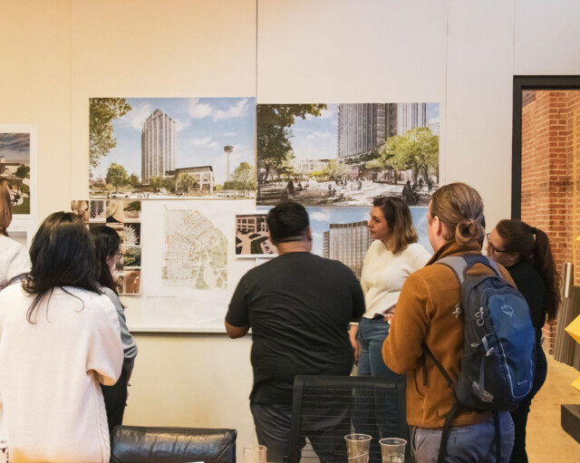 A group of people is gathered in front of a wall with architectural posters, discussing a design project in an office setting, fostering a culture of collaboration and creativity.