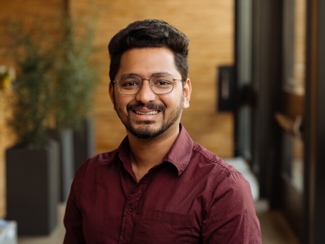 A man with glasses and a maroon shirt, identified as Prathamesh Rewandkar, smiles while standing indoors with blurred background elements.