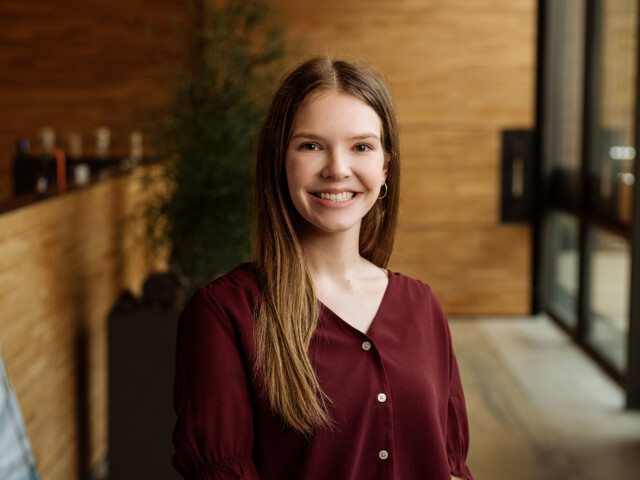 Rachel Kubena, a young woman with long brown hair, is smiling and wearing a burgundy blouse. She stands indoors with a blurred background featuring wooden walls and large windows.