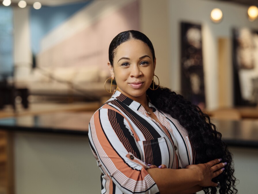 Tori Forte, a woman with long, dark, braided hair and wearing a striped shirt, stands with her arms crossed in a modern, well-lit room.