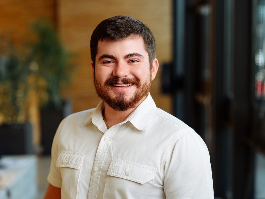 A bearded man, Ty Claussen, smiles while standing indoors. He is wearing a white short-sleeved shirt, and there are plants and large windows in the background.