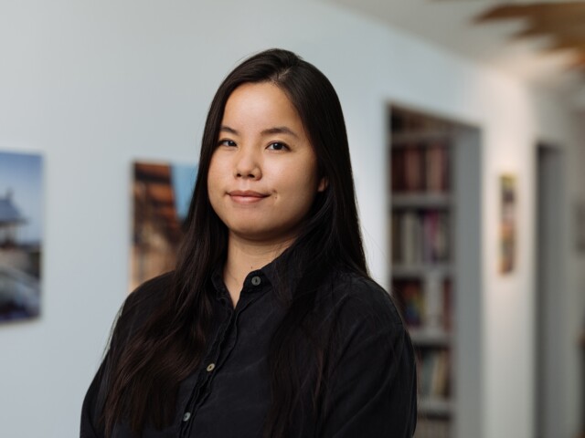 A woman with long dark hair is standing indoors, wearing a black shirt. This headshot of Xiuyin Hu features blurred pictures on the wall behind her and a bookshelf in the background.