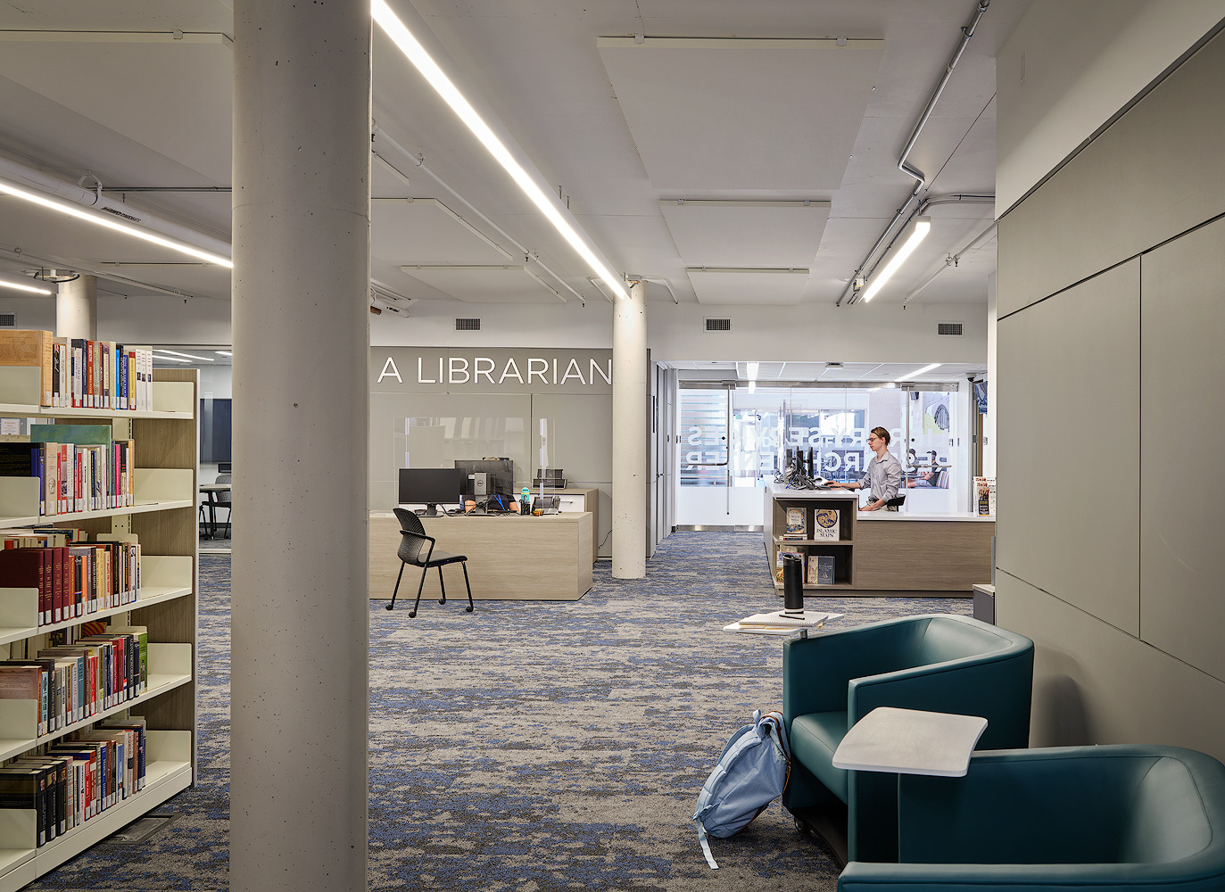 A modern library interior at Austin Community College features bookshelves on the left, a column, a desk labeled "Librarian" in the center, and seating in the foreground. Two people are in the distance near a counter on Rio Grande.