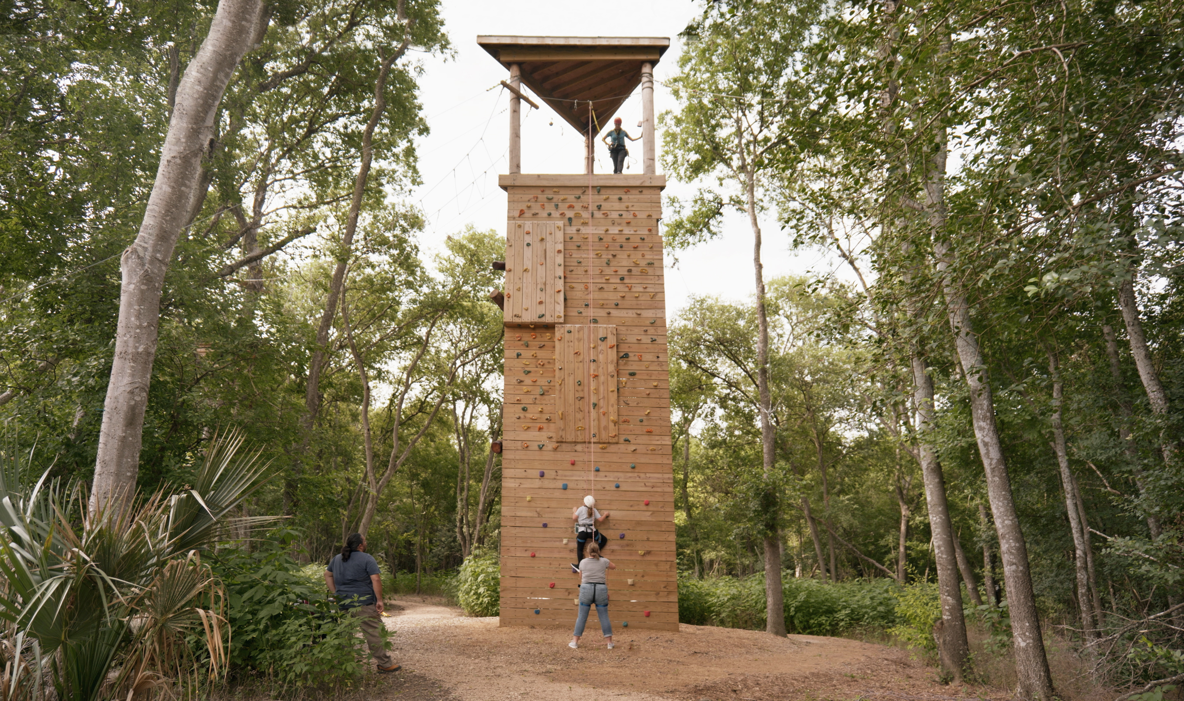 People are engaged in rock climbing and belaying on a tall wooden climbing tower at the Harvey E. Najim Children & Family Center in a forested area, with one person already at the top under a shelter.