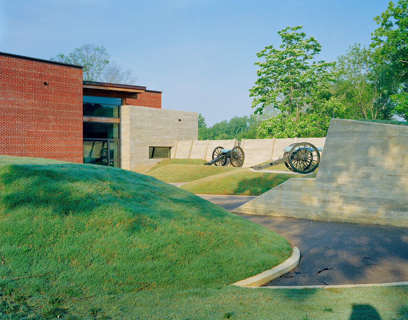 A modern building with a brick façade and concrete walls serves as the Corinth Civil War Interpretive Center, displaying historical cannons on grassy mounds outside, surrounded by trees and an asphalt pathway.